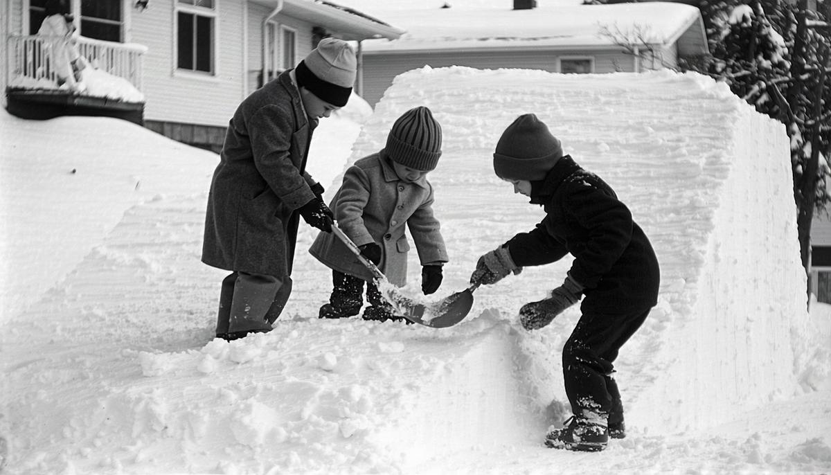 Children in 1950s winter clothing working together to build a snow ramp for sledding
