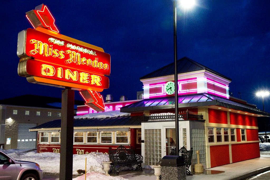 Exterior of a classic 1950s-style diner at dusk, with shiny chrome siding, large windows, and glowing neon signs